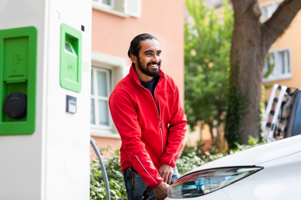 A smiling man in a red jacket plugs in his electric car.