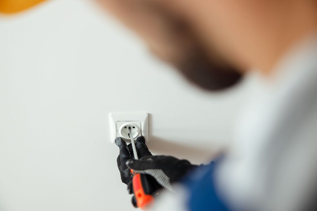 Close-up view of an electrician in protective gloves using a screwdriver to access a socket.