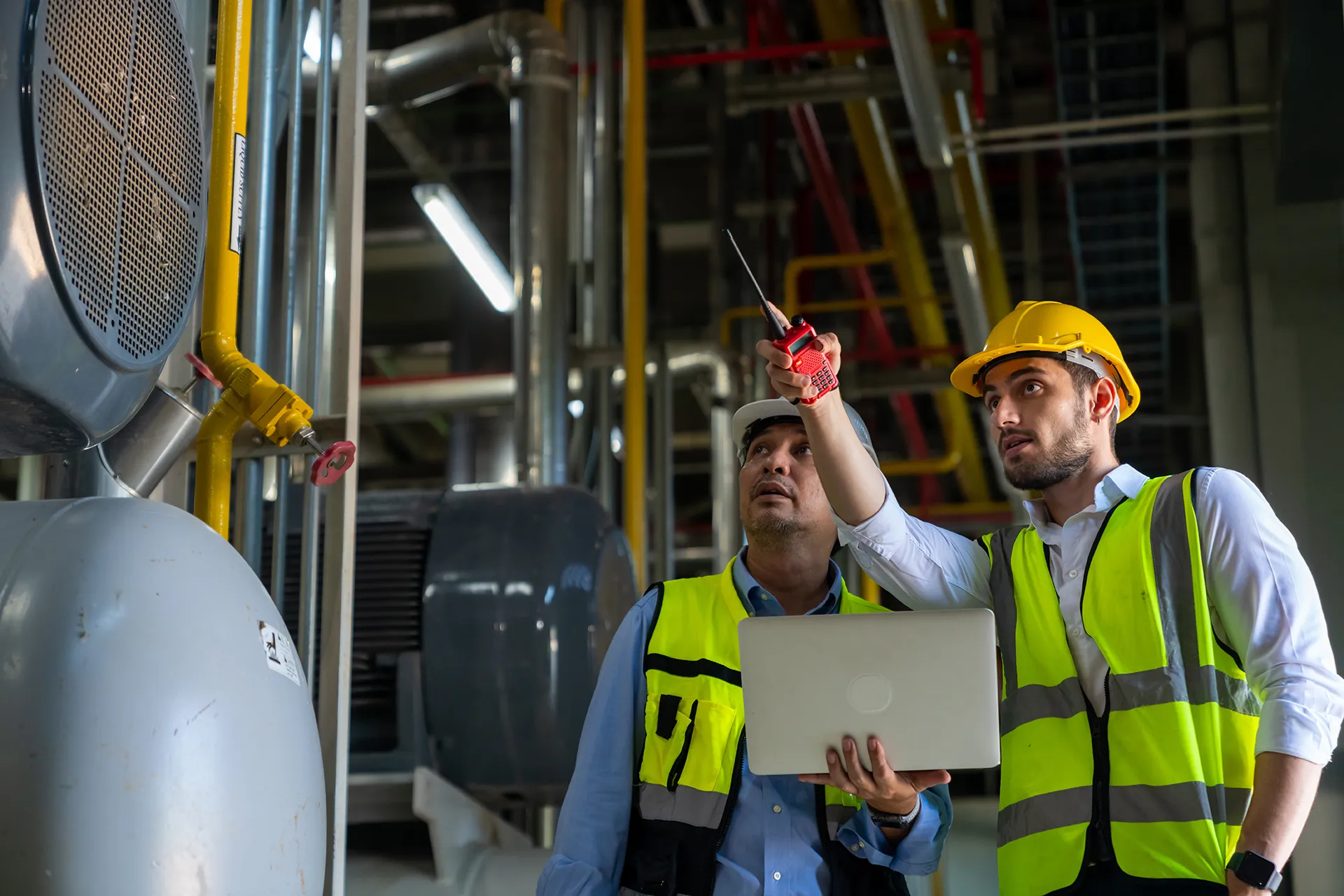 Two male factory workers or engineers in hard hats and safety vests use a laptop and walkie-talkie to coordinate operations.