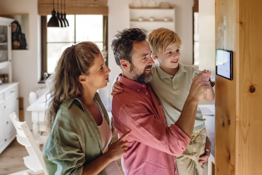 A happy family, a mother, father, and son, stand together as the father and son use a wall-mounted smart home control panel.
