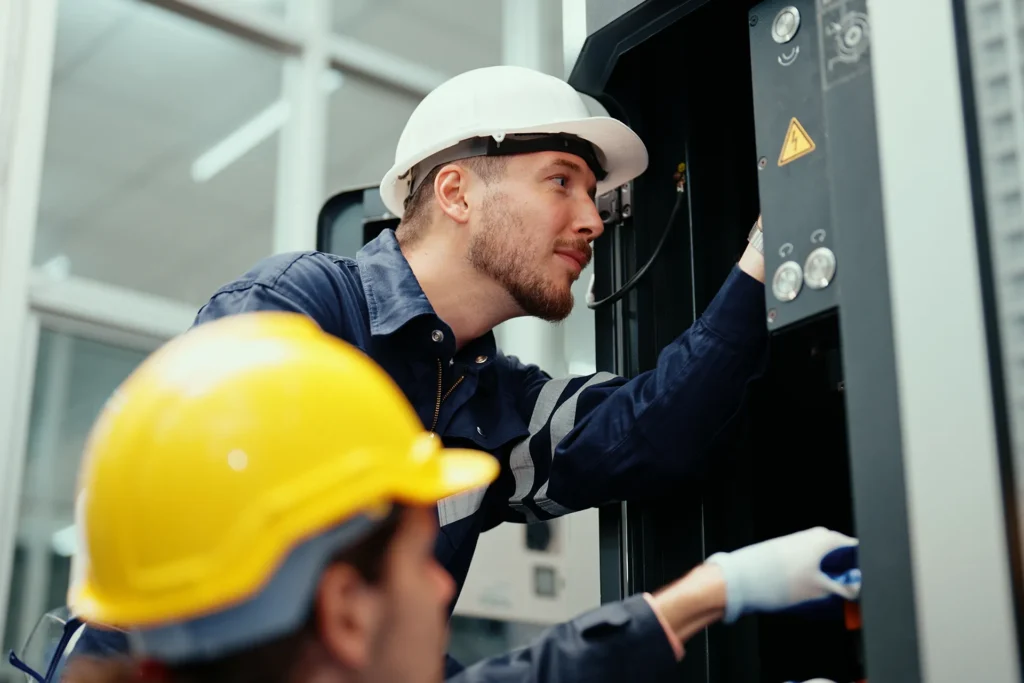 An industrial technician wearing a hard hat and uniform inspects a machine's control panel in a factory.