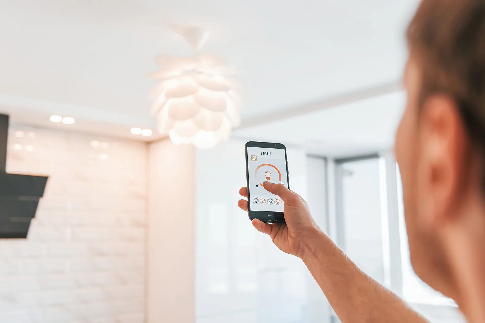 A person uses a smartphone with a smart home app to remotely control a modern light fixture in a room.
