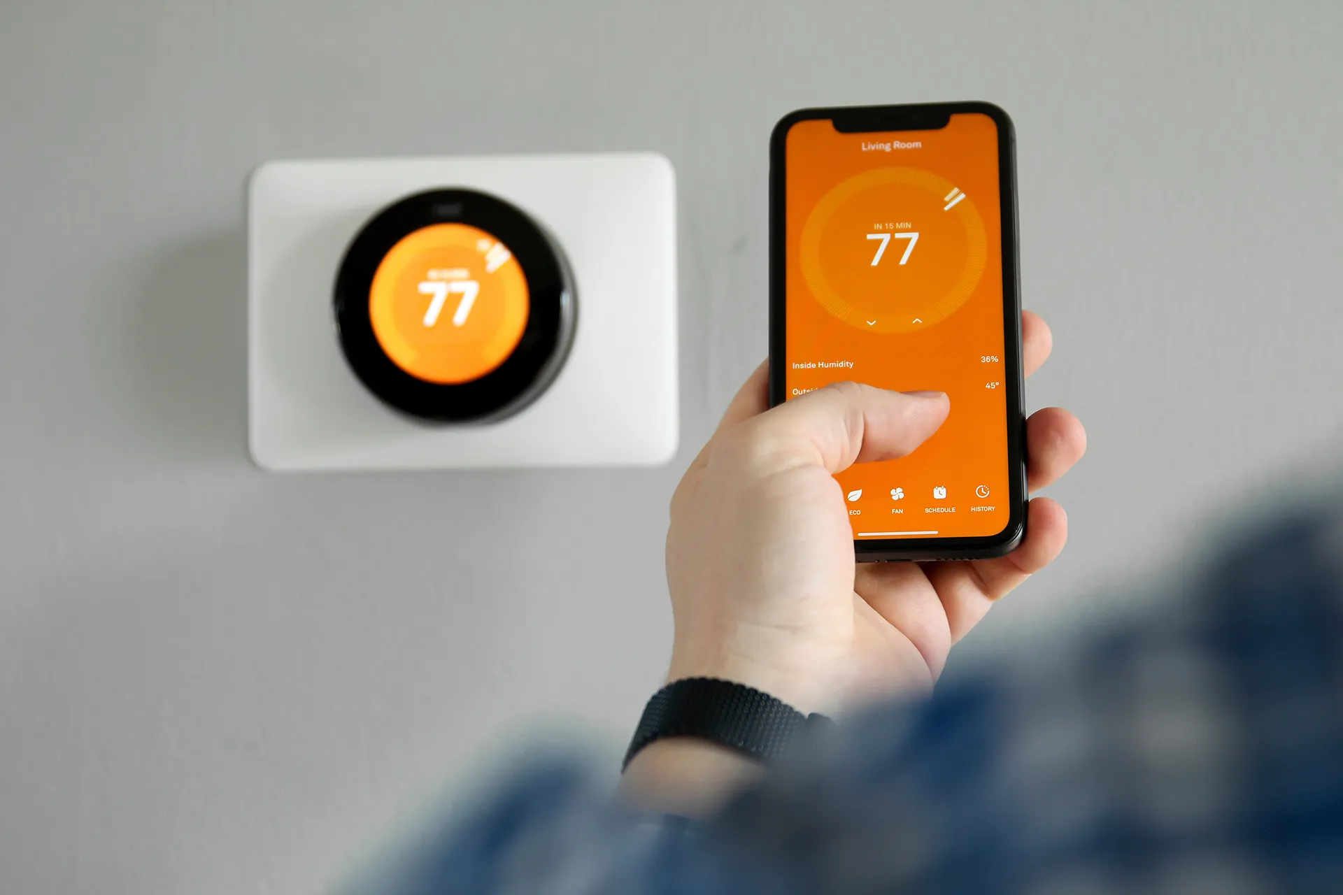 A hand holds a smartphone displaying a smart thermostat app, matching the temperature shown on a wall-mounted smart thermostat.