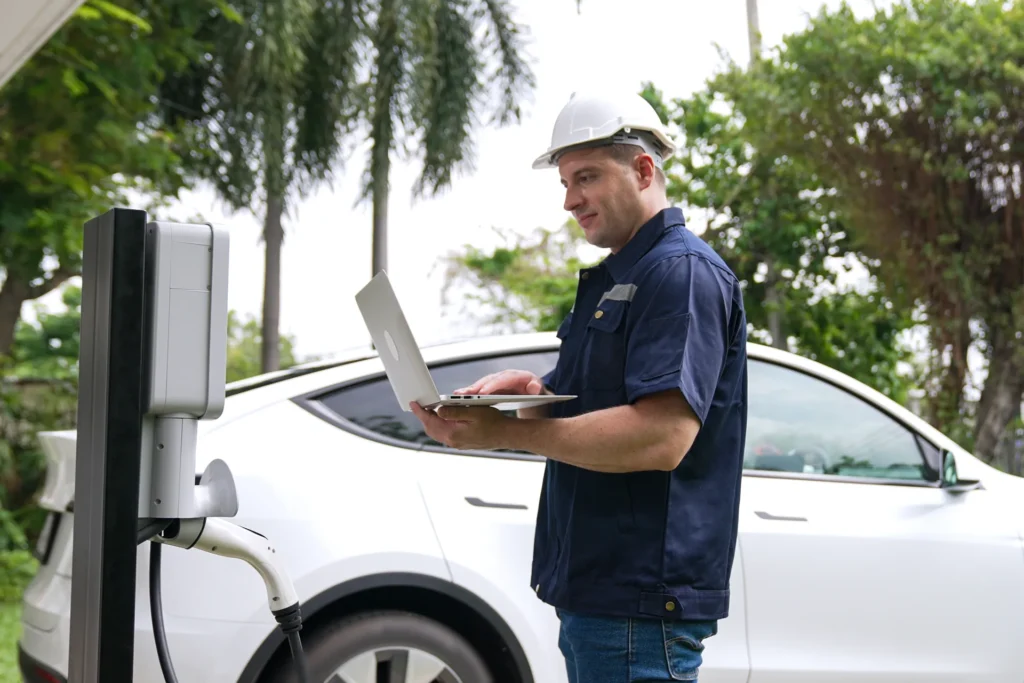 A male technician or electrician in a hard hat uses a laptop to service a residential electric vehicle charging station.