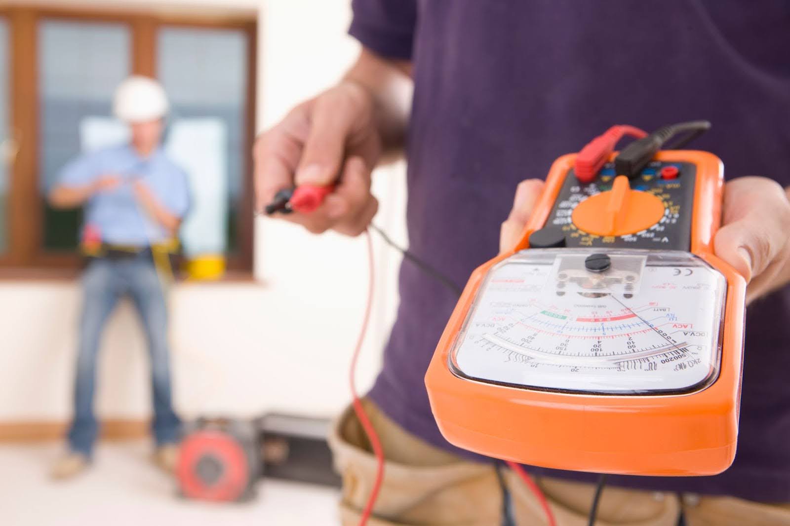 Two electricians working together, one testing voltage with a voltmeter while the other splices a cord.