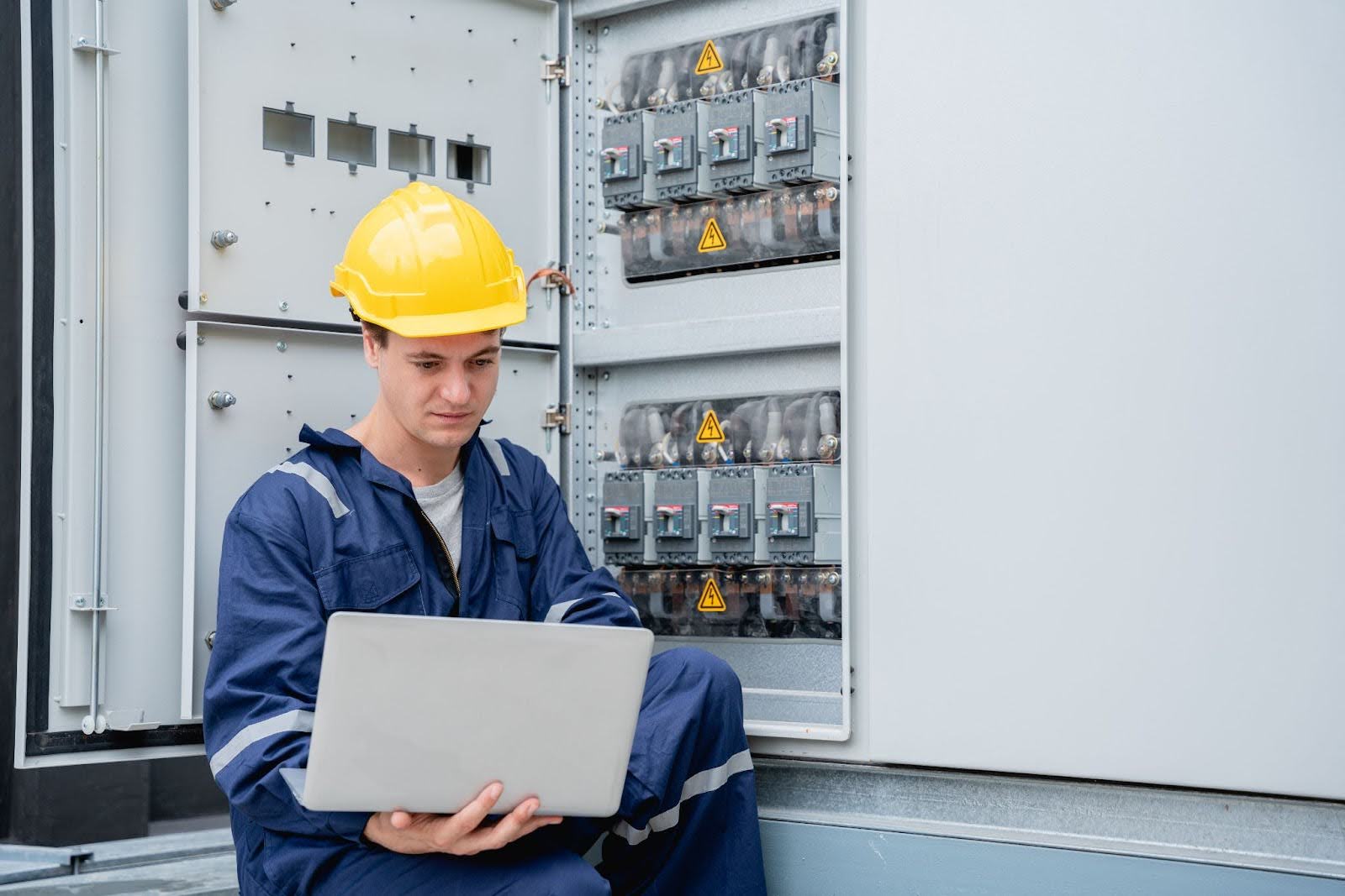 Electrician measuring operating voltage with a laptop at an electrical switchboard during a system inspection.